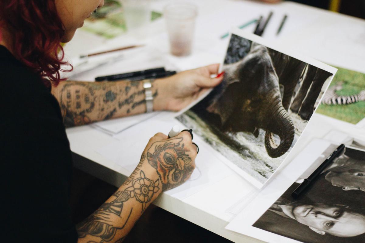 female tattoo artist drawing stencil with reference picture of an elephant