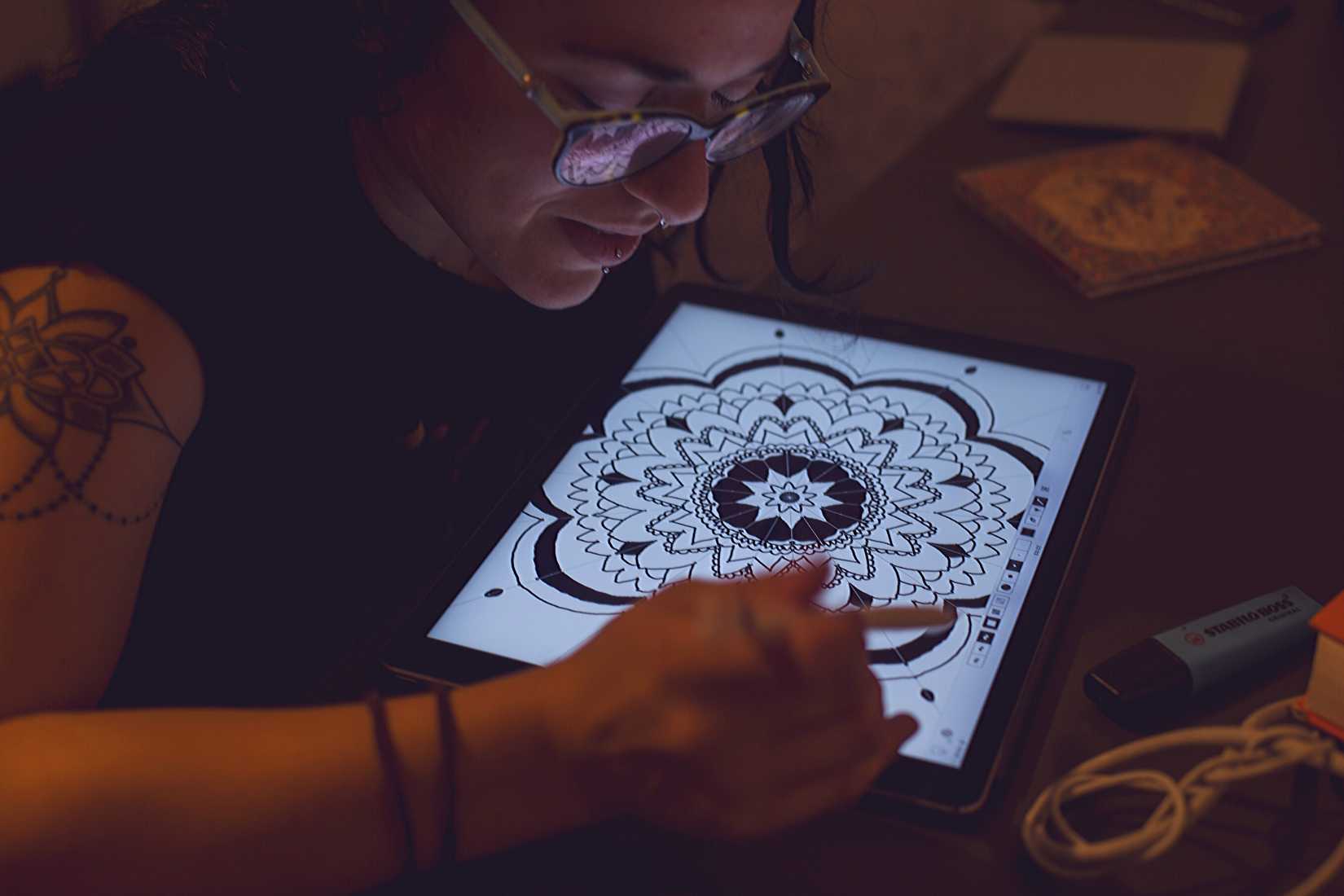 tattooed and pierced woman with glasses using a pent tablet to draw a mandala