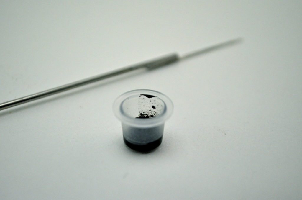 ink cup with black ink and stick and poke needle in the background