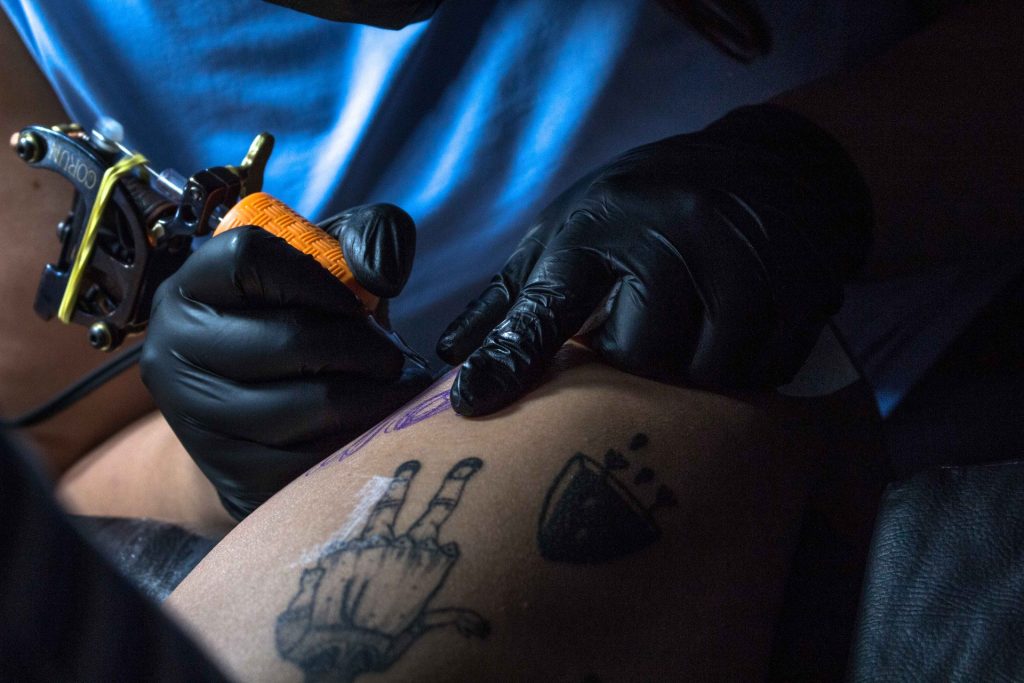 Close-up of an artist using a coil tattoo machine to do line work on a client's thigh. The coil machine is set up with rubber bands, an orange grip, and a metal tube.