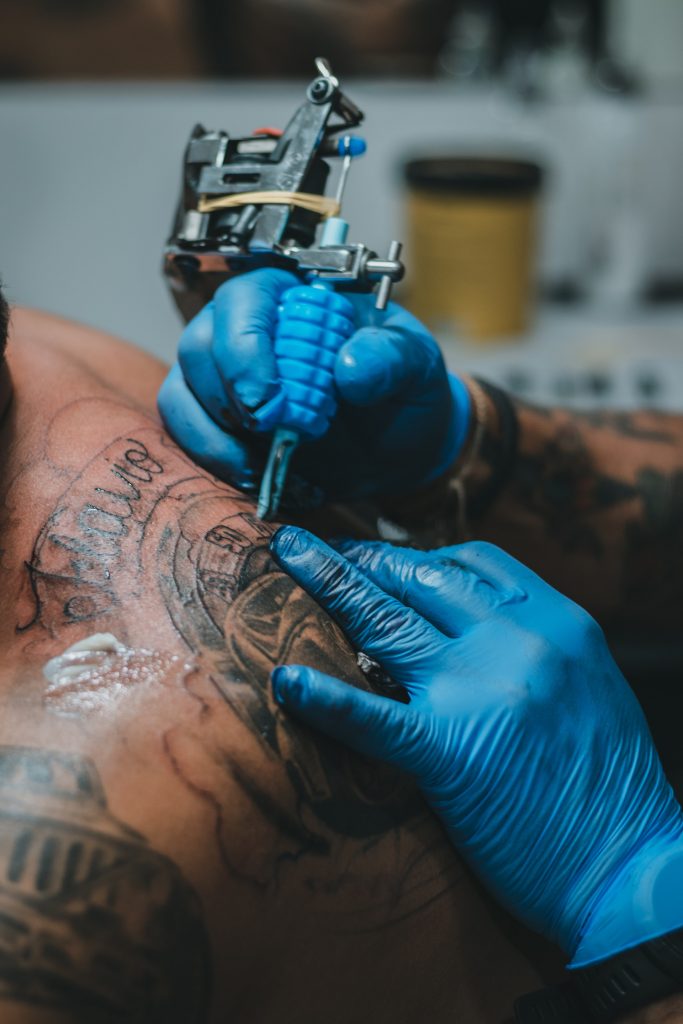 Close-up of an artist using a coil tattoo machine to do line work on a client's back. The coil machine is set up with rubber bands, a textured blue grip, and a metal tube.