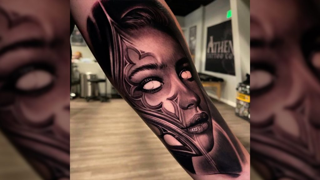 An inner forearm tattoo of a photorealistic, surreal lady head in black and gray. The lady head has all-white eyes and gothic filigree accents on her face.