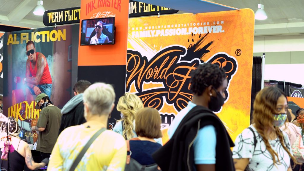 Shot of tattoo convention in progress, including a large banner advertising World Famous Ink with a yellow-and-orange background. Tattoo clients and convention attendants are wandering around looking at the booths.