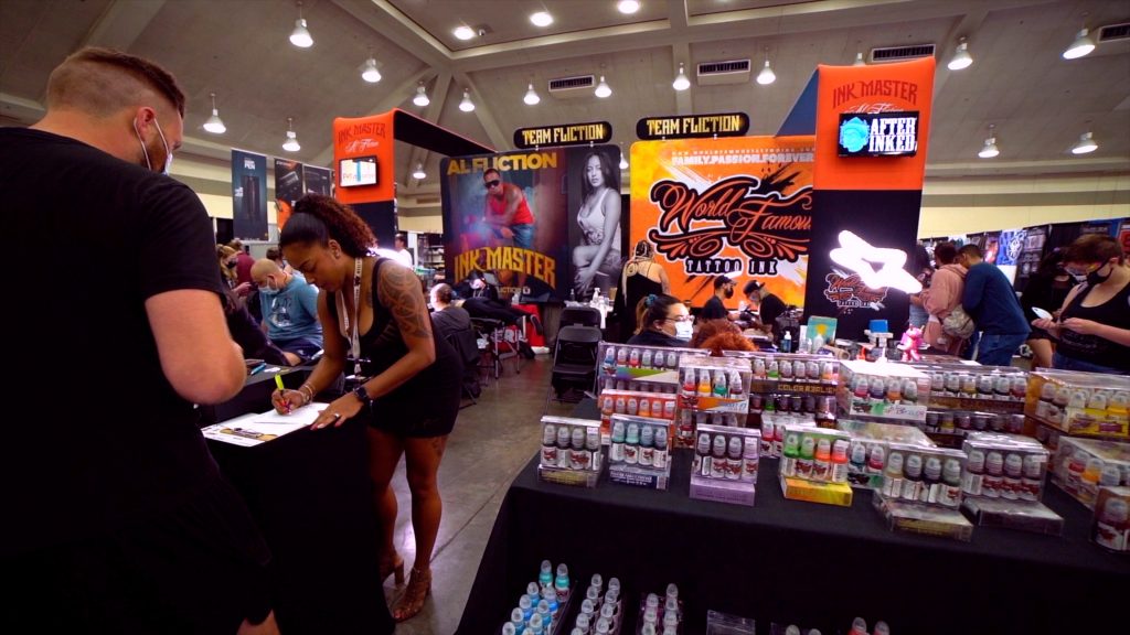Wide shot of tattoo convention featuring several booths hosted by participating vendors, including Ink Master and World Famous Ink. Several boxes of World Famous ink are on display at a booth table.