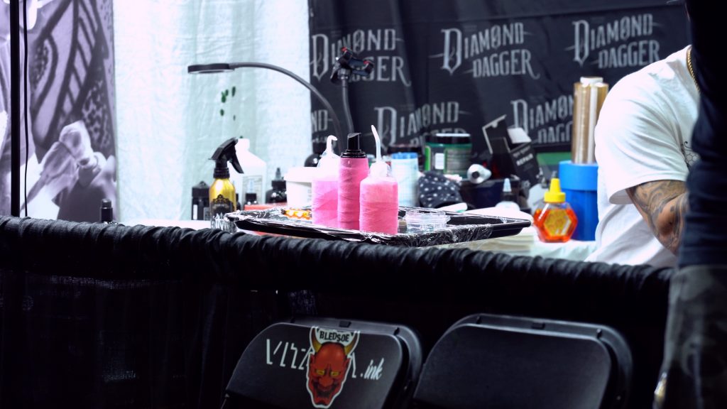 Table at a tattoo convention with a caran-wrapped tray on top of it. The tray includes washer bottles and medical supplies for tattooing. There are also miscellaneous tattoo supplies in the background, including tattoo glide and barrier film. A Diamond Dagger Tattoo curtain is in the backdrop.