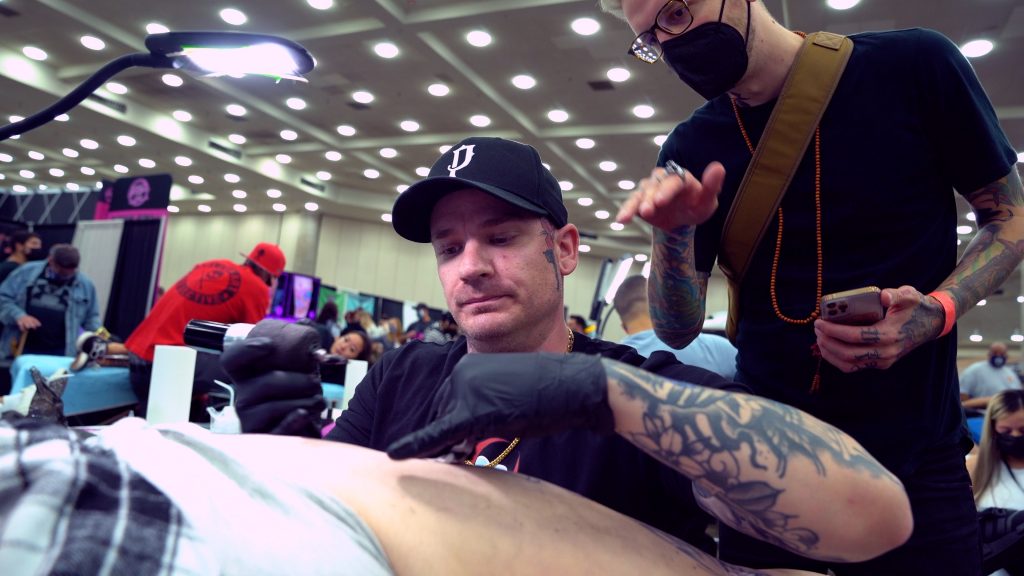 Tattoo artist Harlan Kantner tattooing a client while a fellow artist gives him tips.