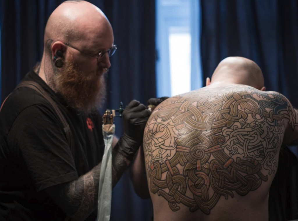 Profile view of Peter Oakmund Madsen tattooing a full back piece on a client. The tattoo has Viking imagery.