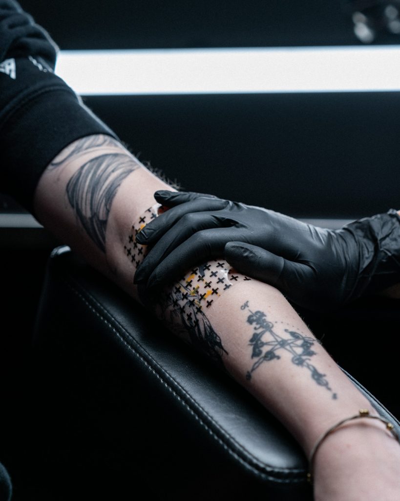 Hand wearing a glove gently pressing on Recovery Derm Shield bandage, which has been applied to tattoo site