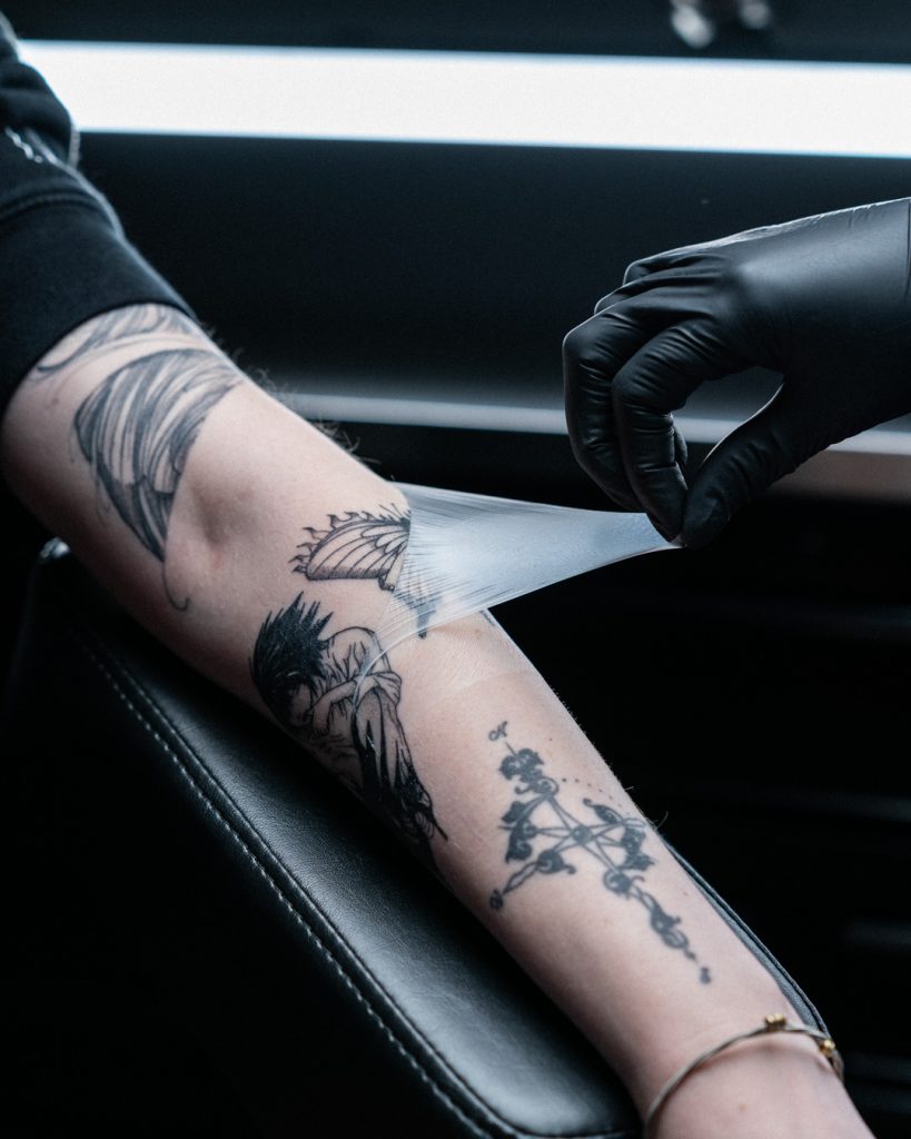 Hand wearing a tattoo glove slowly removing tattoo aftercare bandage from skin, revealing a tattoo of a butterfly