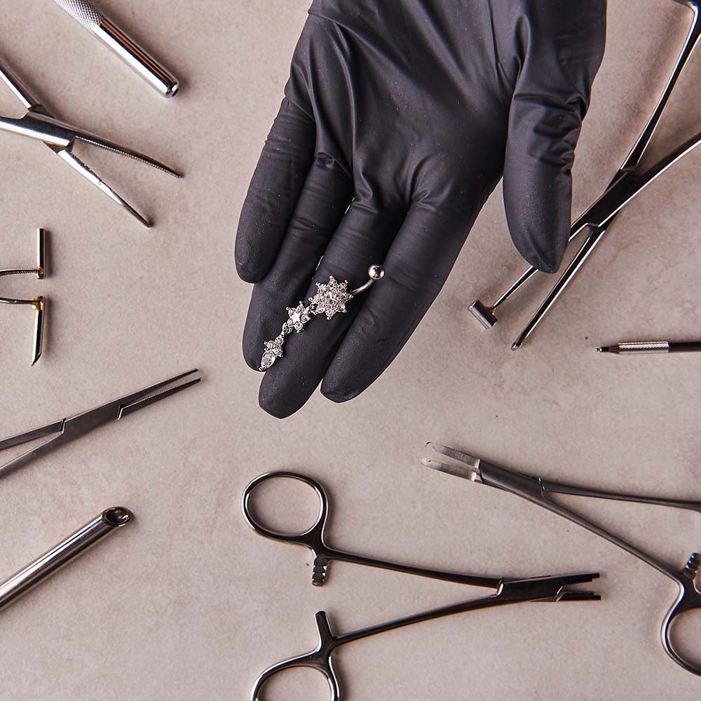 Piercer's gloved hand holding body jewelry over top a table covered in piercing tools, including pliers and forceps